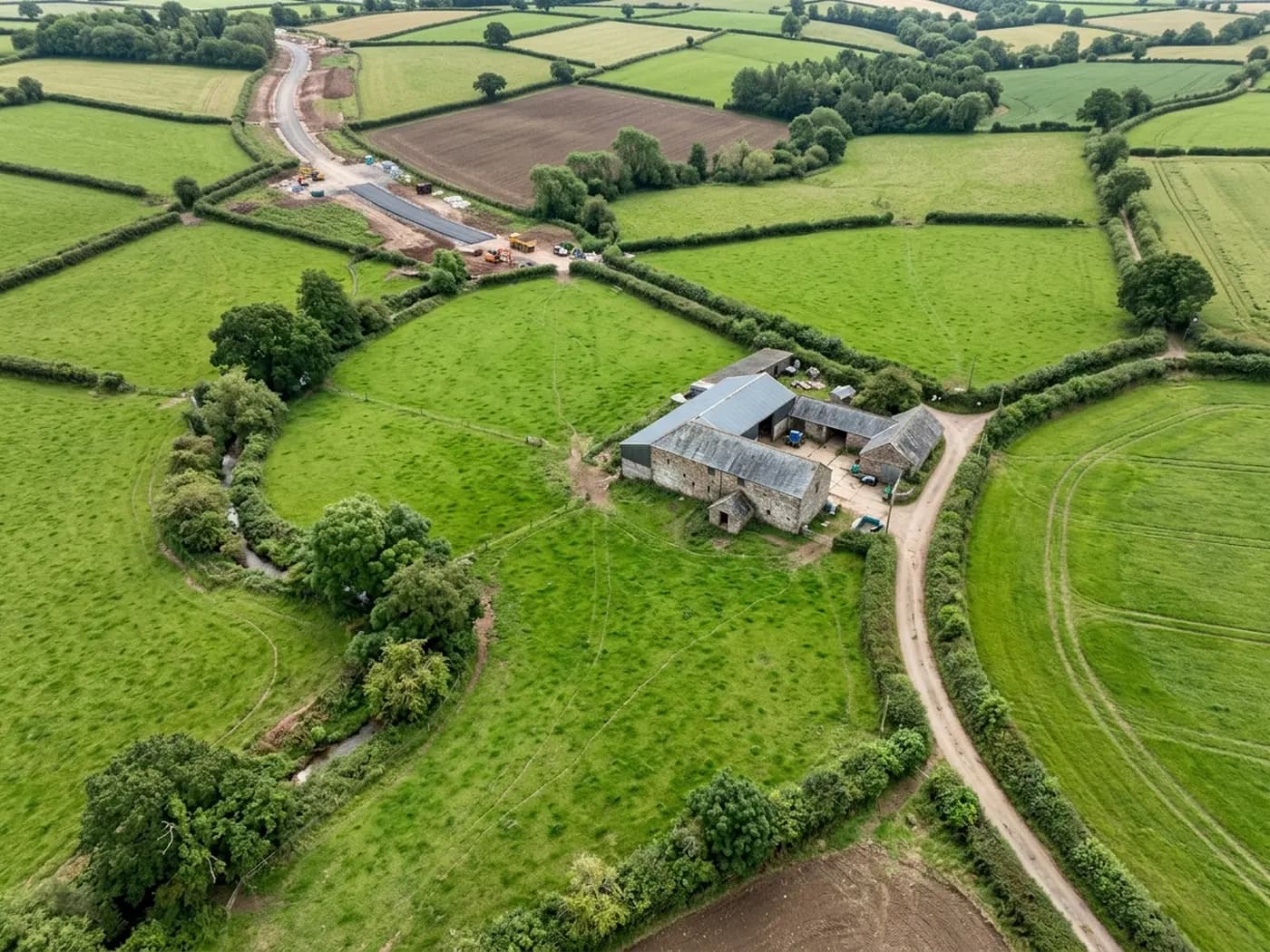 A near-vertical aerial view of Devon farmland showing hedge-banked field boundaries, a farm track, a stone barn and a stream line